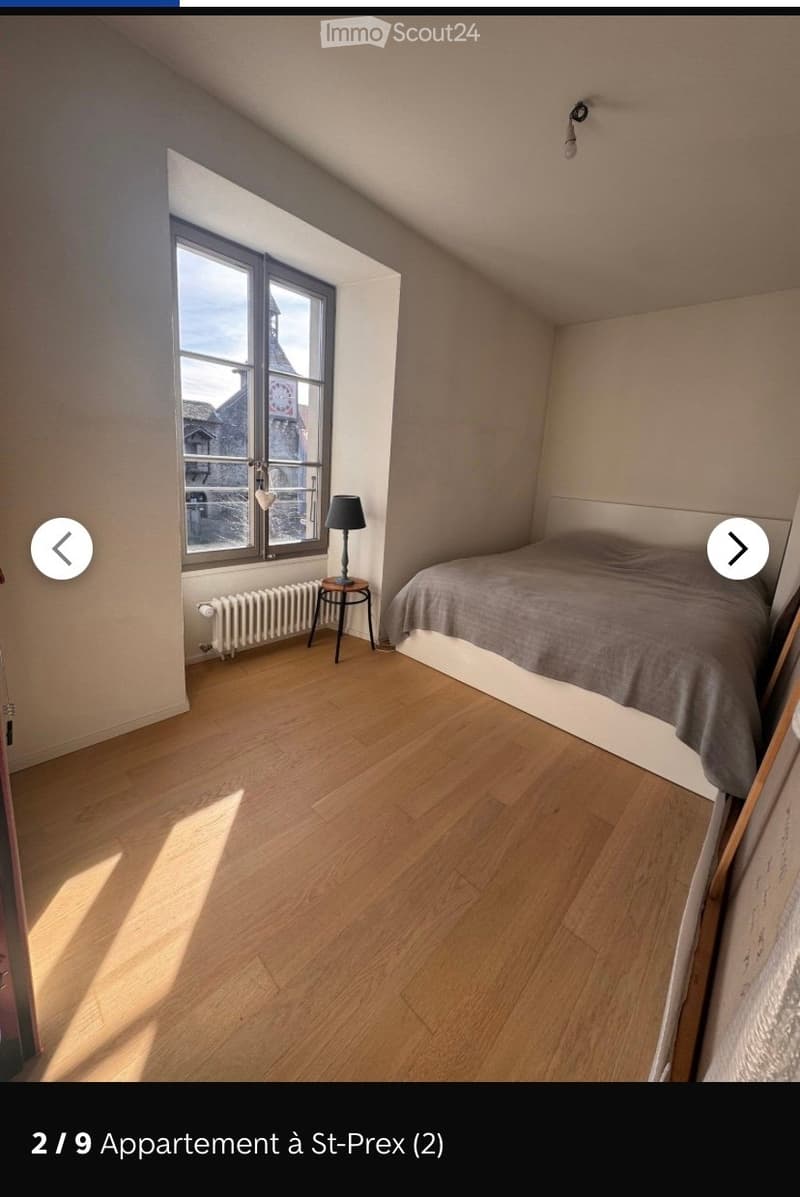 Bright bedroom with wooden flooring, large window offering a view, a single bed with a grey duvet, a small table with a lamp, and a radiator below the window.