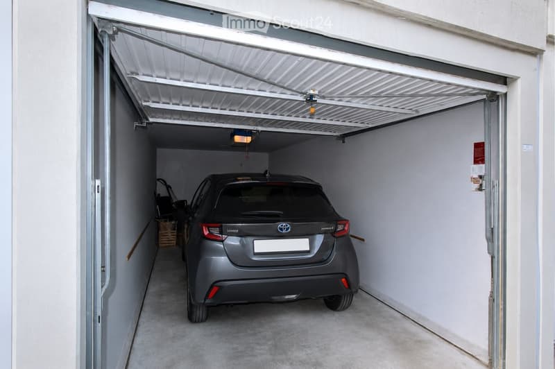 A modern, well-lit garage with a single car parked inside. The garage features a white, automated rolling door, clean white walls, and a concrete floor.