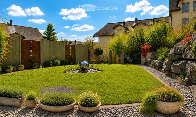 Well-maintained garden with a green lawn, decorative stone pathway, potted plants, and a small water feature. The garden is enclosed by a wooden fence and features a variety of shrubs and trees.