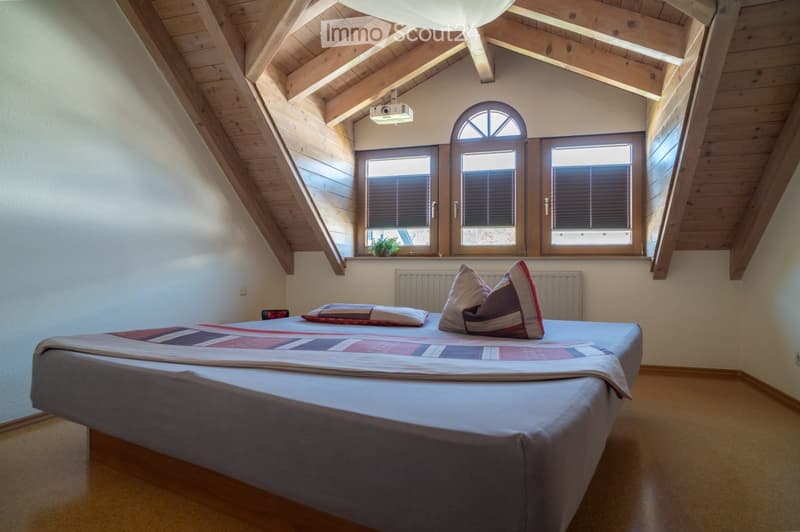 Cozy attic bedroom with wooden slanted ceilings, large windows, a neatly made bed with pillows and a patterned quilt, and a small plant on the windowsill.