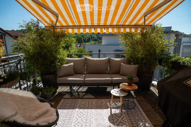 Balcony with a beige sofa, small round table, two stools, a fur throw on a chair, and a patterned rug.
