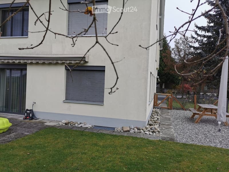 A well-maintained garden area with a lawn, a gravel pathway, a wooden picnic table, and a bench. The house has a light-colored facade with multiple windows and a small awning over the entrance.