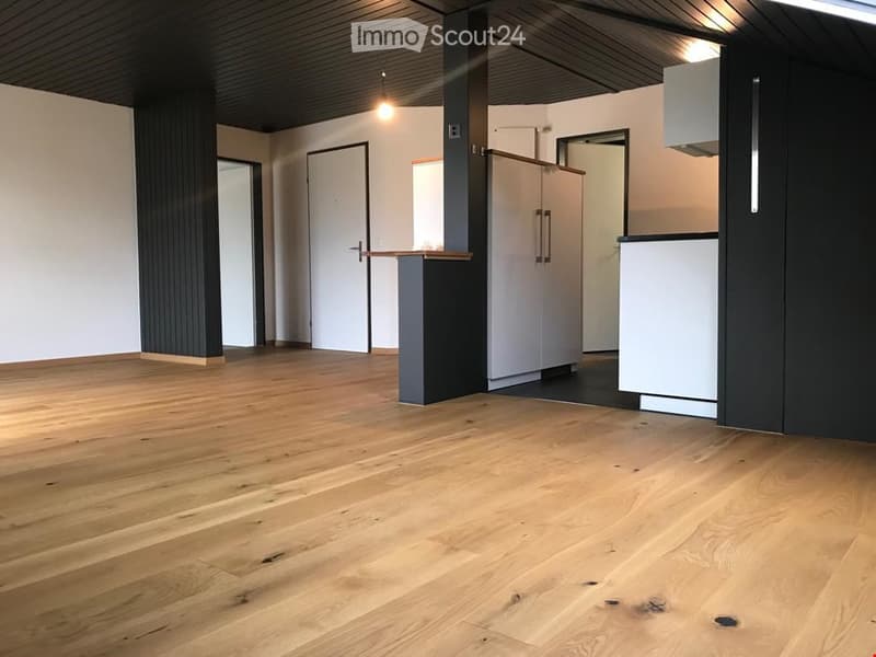 Open-plan space with light wooden flooring, white walls, and modern cabinetry. The area includes a small kitchen section with white cabinets and a countertop, along with a central pillar and several doors leading to other rooms.