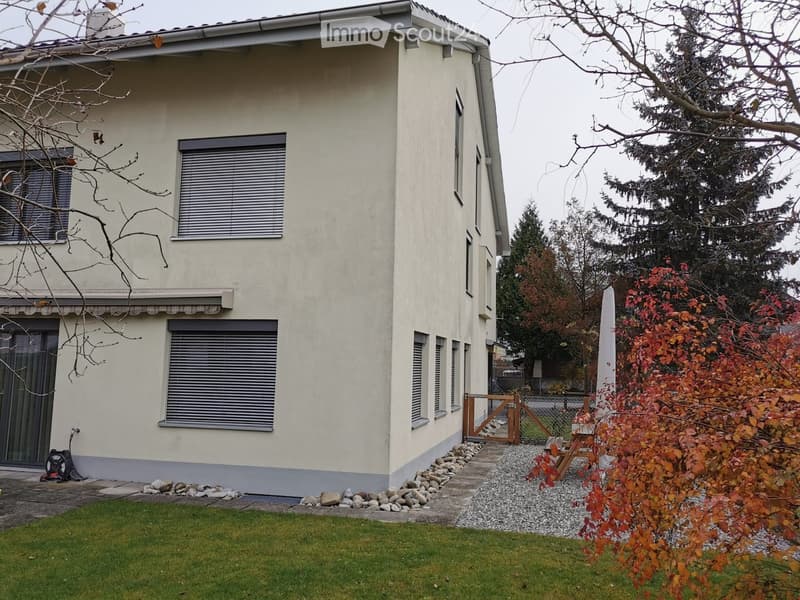 Two-story house with beige exterior, multiple windows with blinds, small garden with grass, gravel pathway, and wooden gate.