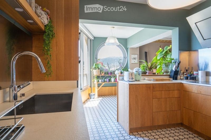 Modern kitchen with wooden cabinets, a large window offering a scenic view, a sink with a modern faucet, and various appliances including a blender and toaster. Potted plants are placed on a shelf near the window.