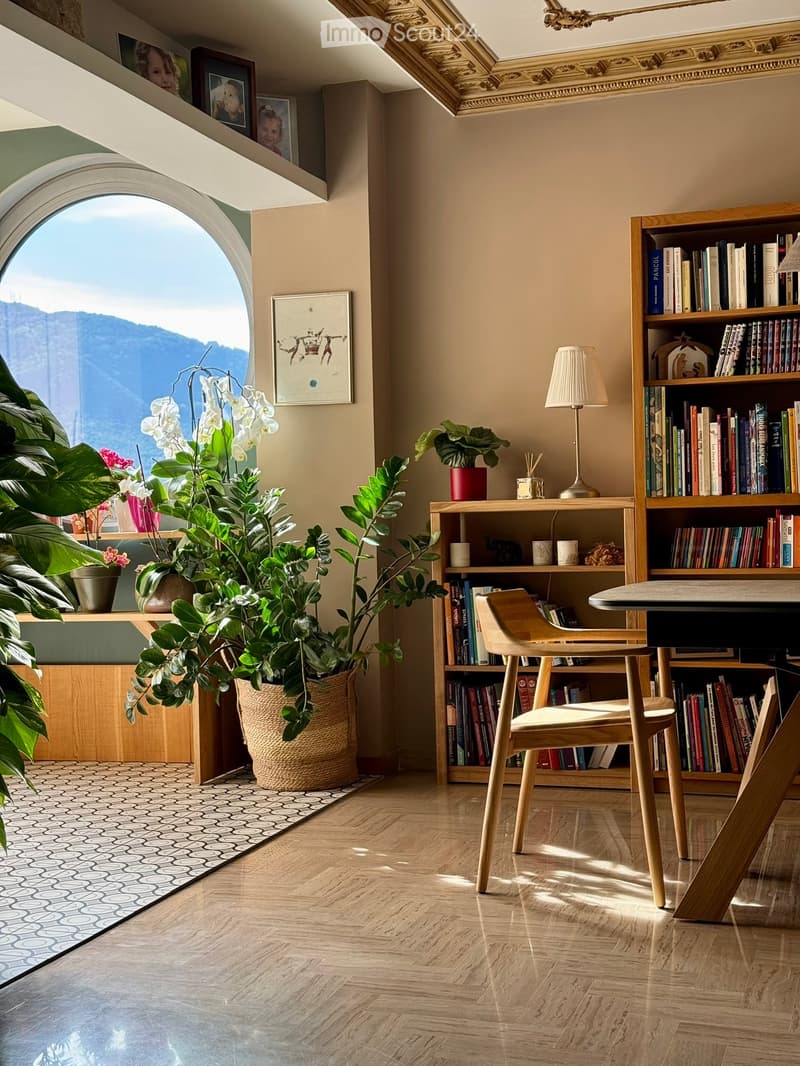 Bright, modern room with wooden flooring, large arched window offering a scenic view, bookshelf filled with books, a small table with a lamp, and several potted plants.