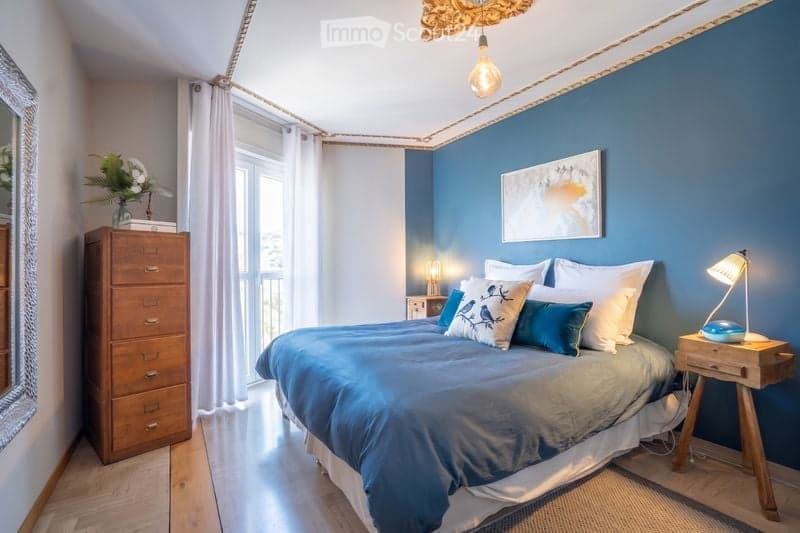 Cozy bedroom with blue walls, wooden floor, large bed with blue and white bedding, nightstands with lamps, dresser, mirror, and decorative vase on the dresser.