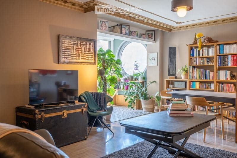 A cozy living room with a large window, green plants, a bookshelf filled with books, a coffee table, a sofa, and a television. The room features warm lighting and a mix of modern and classic decor.