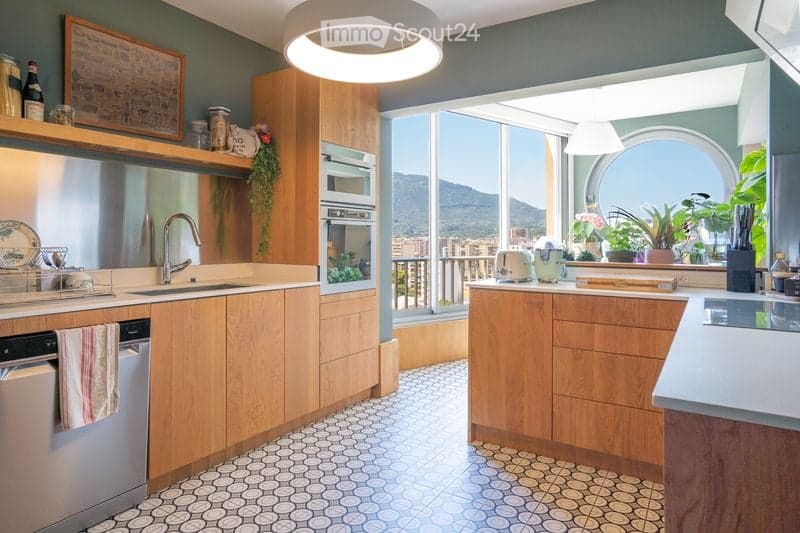 Modern kitchen with wooden cabinets, stainless steel appliances, and a view of the mountains through large windows. Features include a sink, oven, dishwasher, and various kitchen items on the counter.