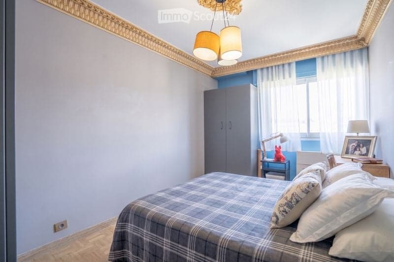 A modern bedroom with a grey plaid bedspread, white pillows, a grey wardrobe, a small table lamp, and a framed picture on a nightstand.
