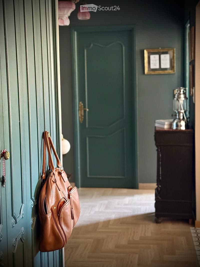 A hallway with dark green walls, wooden flooring, and a leather bag hanging on the door. A small table with a lamp and books is visible in the background.