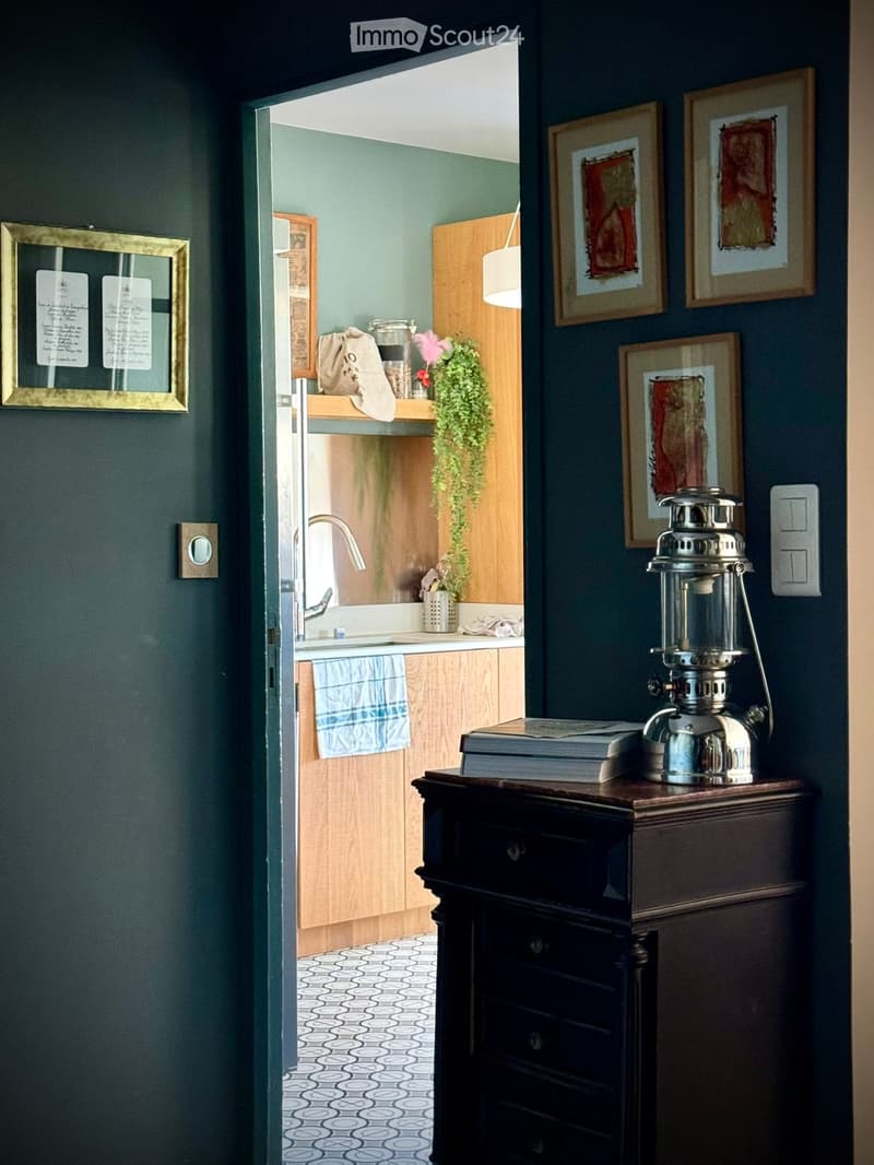 A hallway with dark green walls leads to a small kitchen area. The hallway features a wooden table with a lantern and books, framed pictures on the walls, and a patterned floor. The kitchen has wooden cabinets, a sink, a towel, and a small plant on the co