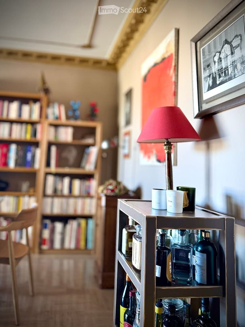 A cozy hallway with a wooden bookshelf filled with books, a small table with a lamp and decorative cups, and a wooden chair.