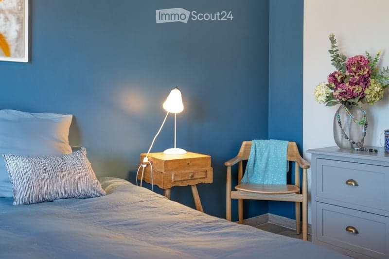 Cozy bedroom with blue walls, a neatly made bed, a wooden nightstand with a lamp, a small wooden chair with a blue towel, and a decorative vase with flowers on a dresser.