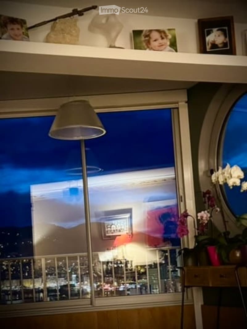 A cozy living space with large windows offering a city view, a floor lamp, a small table with a flower pot, and decorative items on the shelf above the window.