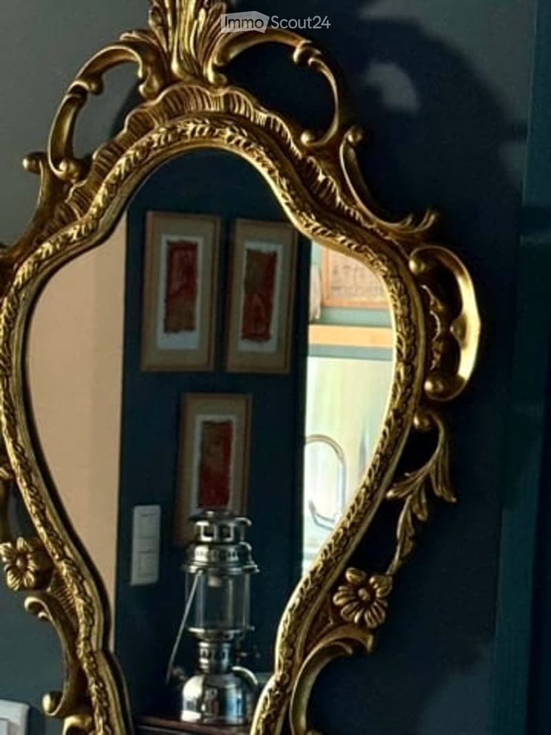 An ornate gold-framed mirror with a decorative design, reflecting a lantern and picture frames on a dark blue wall.