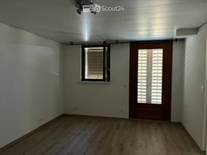 Empty room with wooden flooring, white walls, a single window with blinds, and a wooden door with louvered shutters.