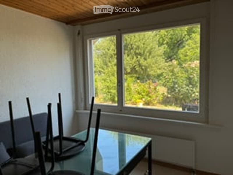 A small, unfurnished room with a large window offering a view of greenery outside. A single table and a chair are placed near the window.