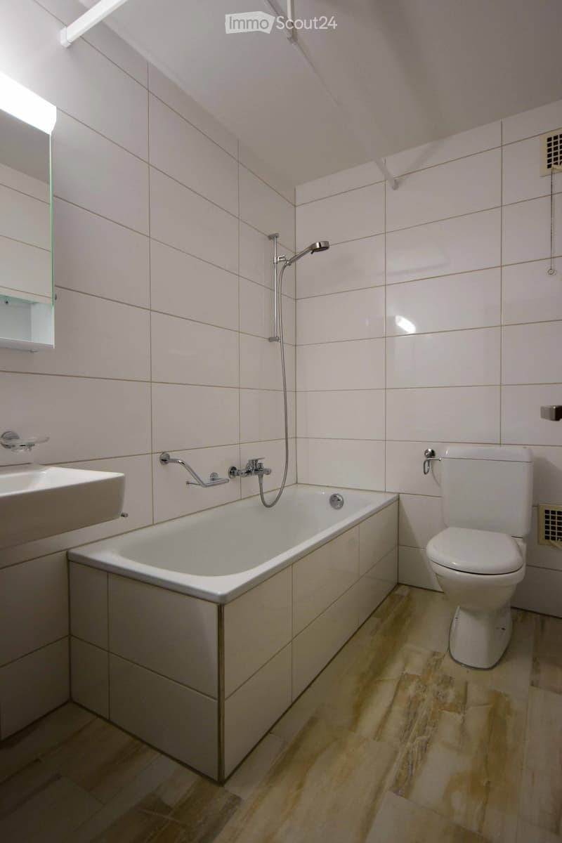 Modern bathroom with white tiles, a bathtub, toilet, sink, and a mirror. The floor is made of light-colored wood.