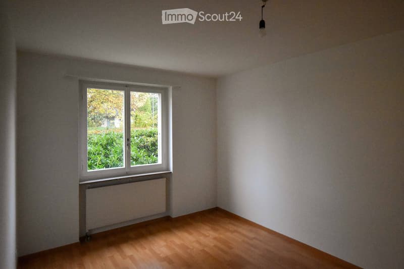 Empty room with wooden flooring, white walls, a single window with white frame, radiator below the window, and a ceiling light fixture.
