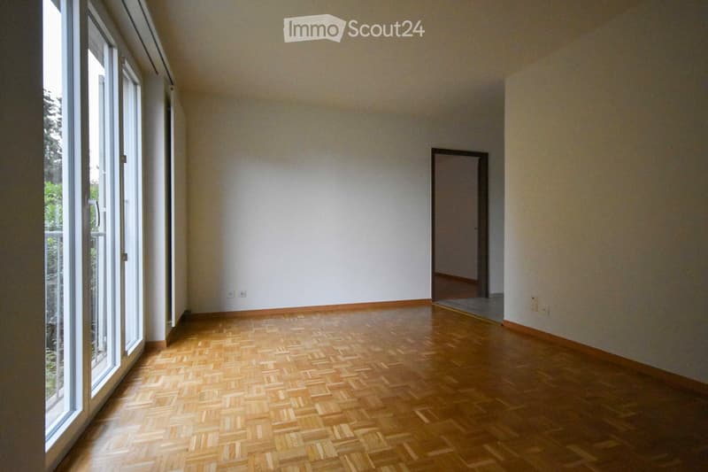 Empty room with wooden parquet flooring, white walls, large window with white frame, and a doorway leading to another space.