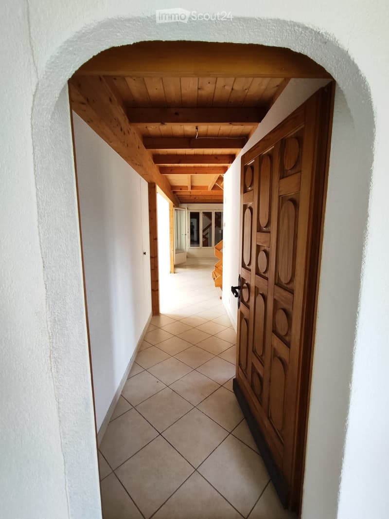 A long, narrow hallway with white walls, tiled floor, and wooden ceiling beams. Features a large wooden door on the right and an open doorway at the end leading to another area.