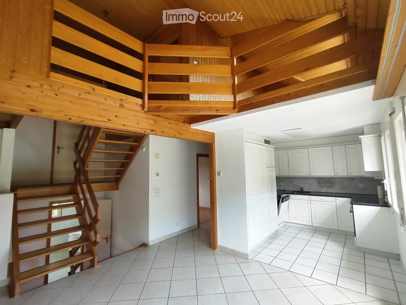 The image shows a modern, well-lit interior space with a wooden staircase leading to an upper level, a kitchen area with white cabinets, a sink, and an oven, and tiled flooring throughout.