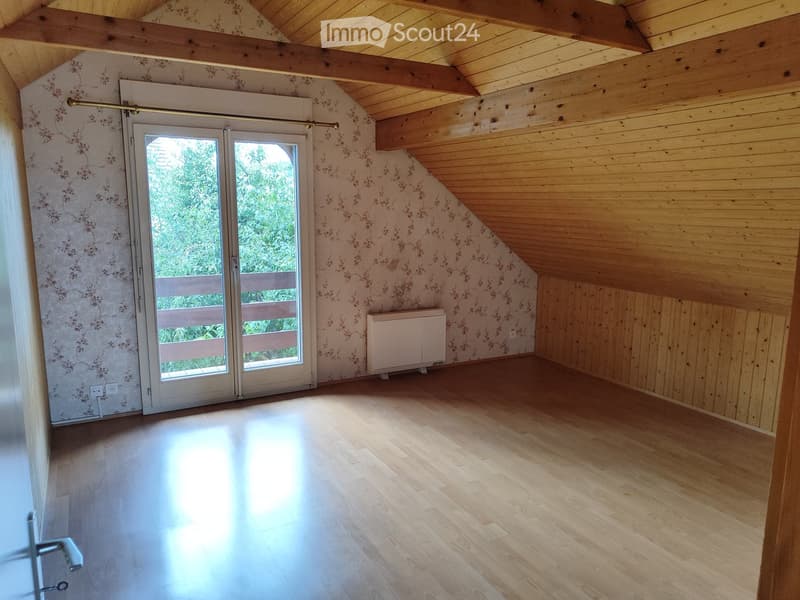 Empty attic room with wooden floors, exposed wooden beams, floral wallpaper, and a large window with a view of greenery.