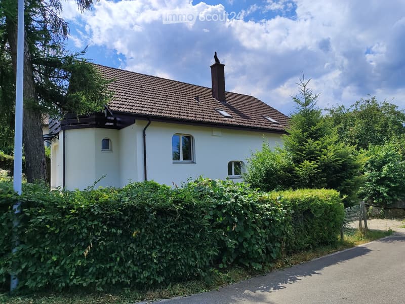 Single-story house with a tiled roof, white exterior, and small chimney. Surrounded by greenery and a hedge.