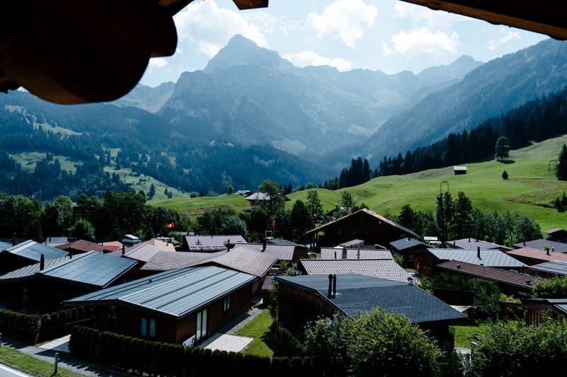 A picturesque village nestled in a lush green valley with snow-capped mountains in the background. Traditional wooden houses with pitched roofs are scattered across the landscape, surrounded by trees and open fields.