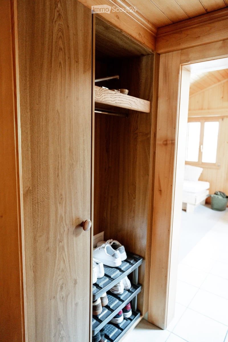 A wooden wardrobe with open shelves and a shoe rack inside, featuring neatly arranged shoes and other items. The room has light wooden walls and a tiled floor.
