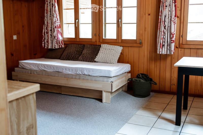 Cozy bedroom with wooden walls, a simple wooden bed with pillows, a small table, and a basket on the floor.