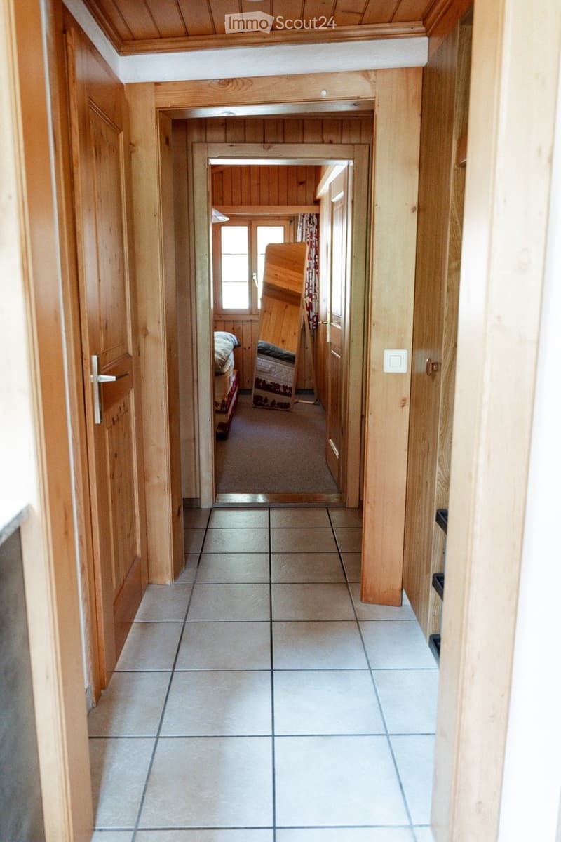 A narrow hallway with wooden walls and tiled floor, leading to a bedroom area with a bed and wooden furniture.