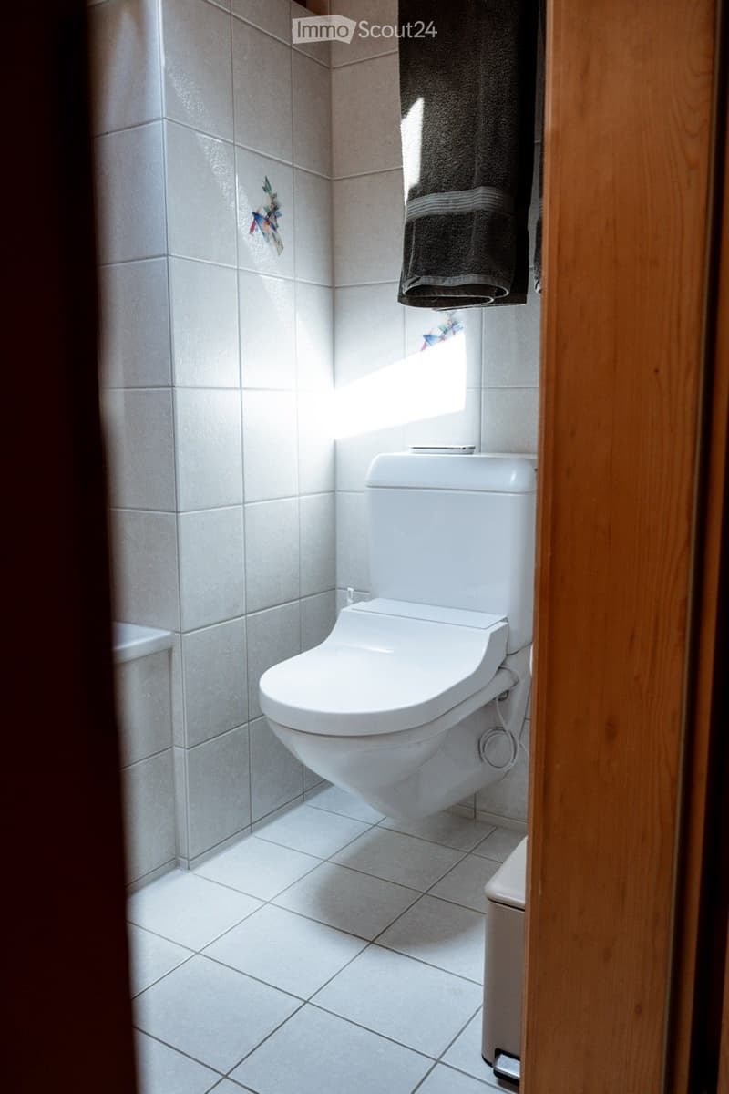 Modern bathroom with white tiled walls and floor, a toilet, and a towel hanging on the door.