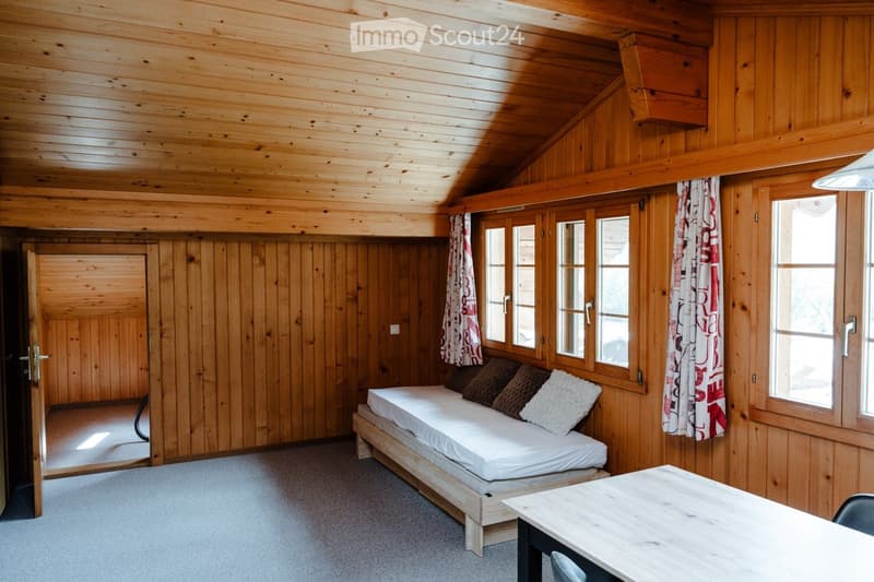 Cozy wooden room with a daybed, throw pillows, a small table, and chairs. The room features wooden walls, a slanted ceiling, and large windows with curtains.