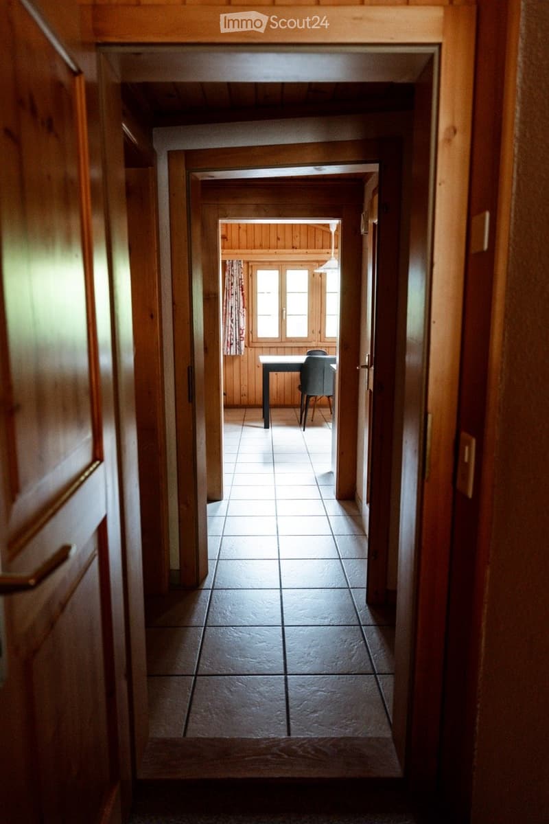 A long, narrow hallway with wooden walls and tiled flooring, leading to a room with a table and chair visible at the end.