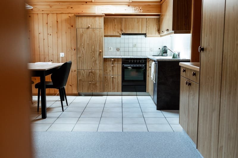 Modern kitchen with wooden cabinets, tiled backsplash, and a black oven. Includes a dining table and chair.