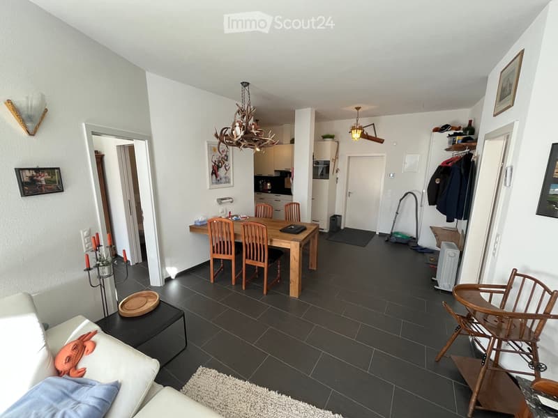 Open-plan living and dining area with a white sofa, wooden dining table, chairs, baby chair, and a chandelier. The kitchen is visible in the background with cabinets and appliances.