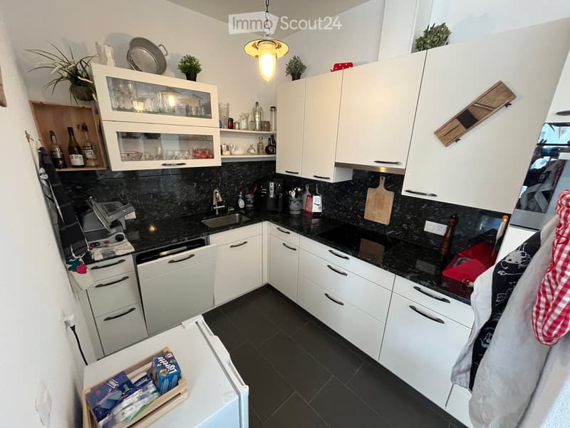 Modern kitchen with white cabinets, black countertops, and various appliances including a dishwasher, coffee maker, and cutting board. The space features a sink, faucet, and a small refrigerator with a wooden tray on top.