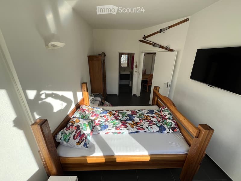 Cozy bedroom with a wooden bed, colorful butterfly-patterned bedding, a wardrobe, a television mounted on the wall, and a nightstand with a lamp. The room has white walls and a dark tiled floor.