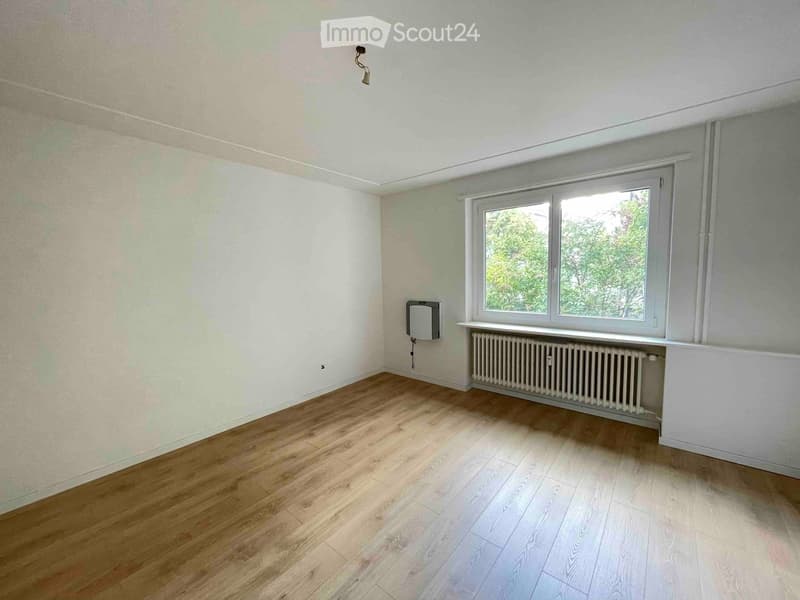 Empty room with wooden flooring, white walls, and a large window with a radiator below it. No furniture or personal items are present.