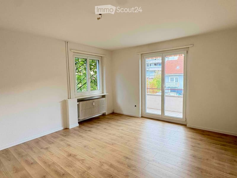 Empty room with light wooden flooring, white walls, a radiator, and large windows with a sliding glass door leading to a balcony.