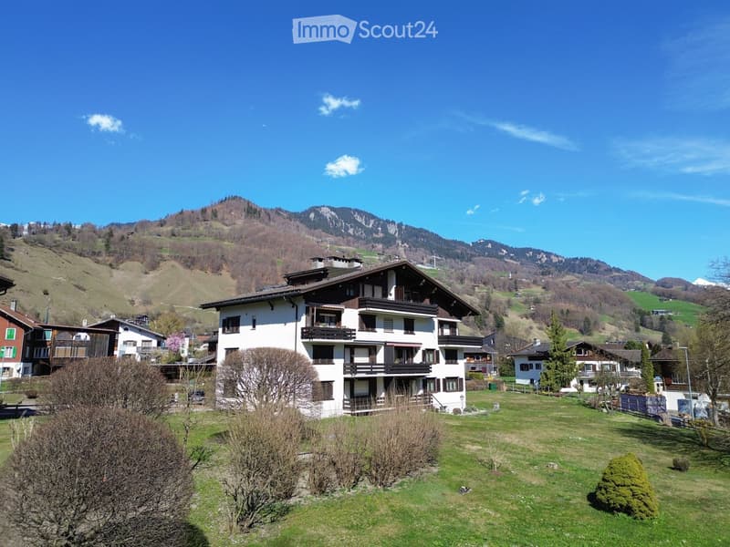 A multi-story house with balconies and a well-maintained lawn, surrounded by other houses and a scenic mountain backdrop.