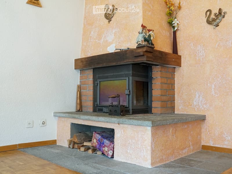 A cozy living room with a brick fireplace, wooden mantel, decorative gnome figurines, a vase with flowers, and a stack of firewood nearby.