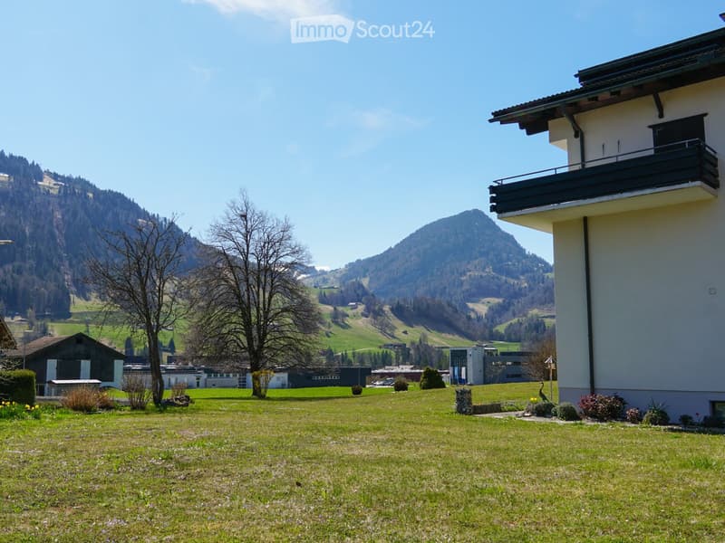 A modern house with a balcony overlooking a green lawn and scenic mountain backdrop.