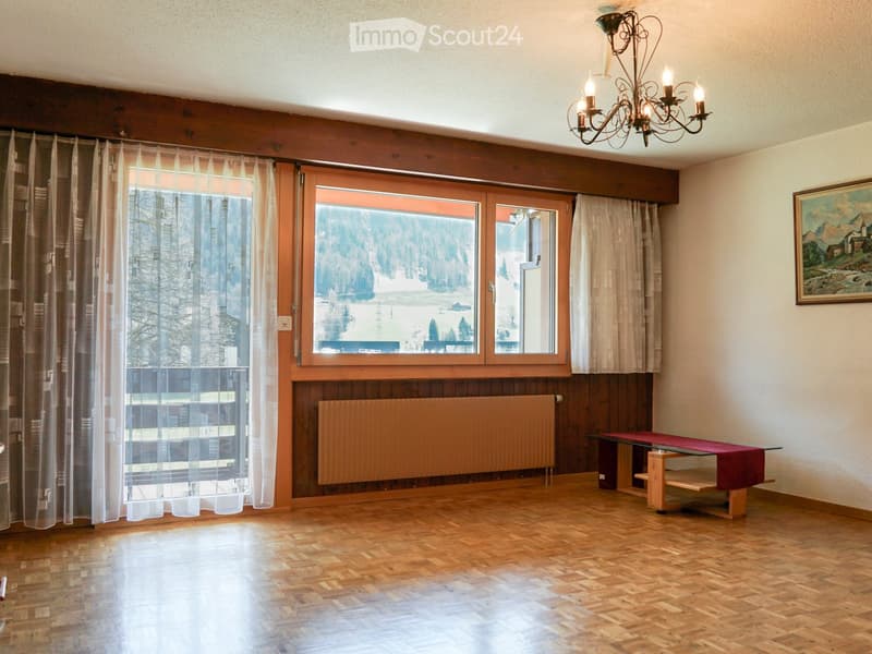 Bright room with wooden flooring, large windows with sheer curtains, a radiator, a small table with a red cloth, a chandelier, and a painting on the wall.