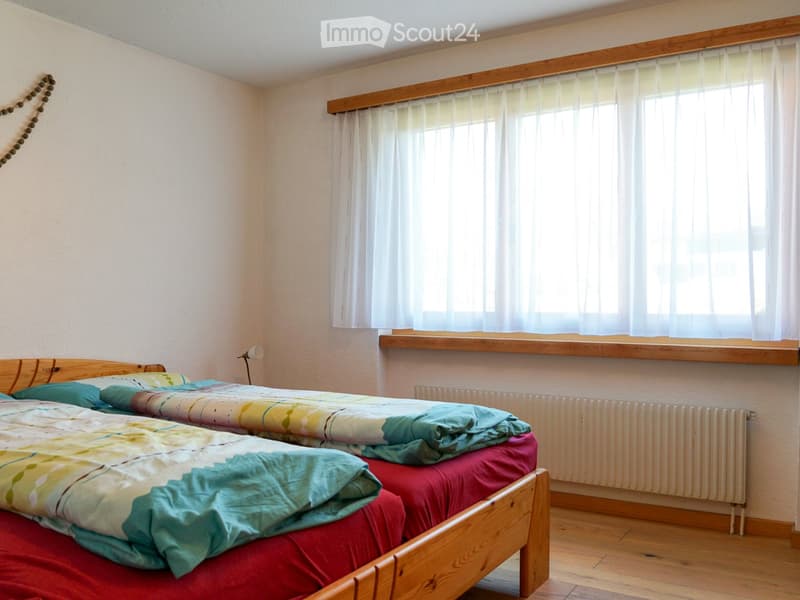 Cozy bedroom with wooden floors, two single beds with colorful bedding, a radiator, and a window with sheer curtains.