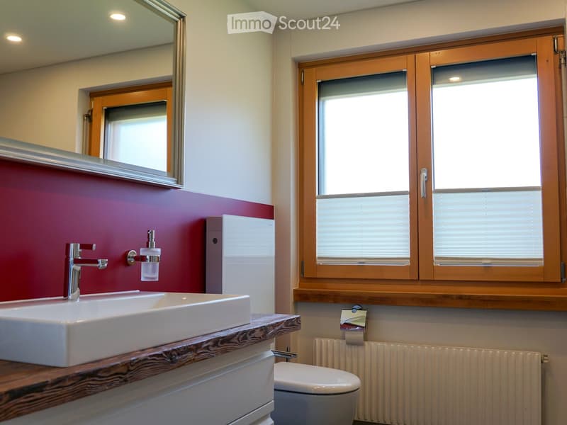 Modern bathroom with a white sink, chrome faucet, toilet, large mirror, and wooden-framed windows with blinds. The walls are painted in a two-tone design with red and white sections.