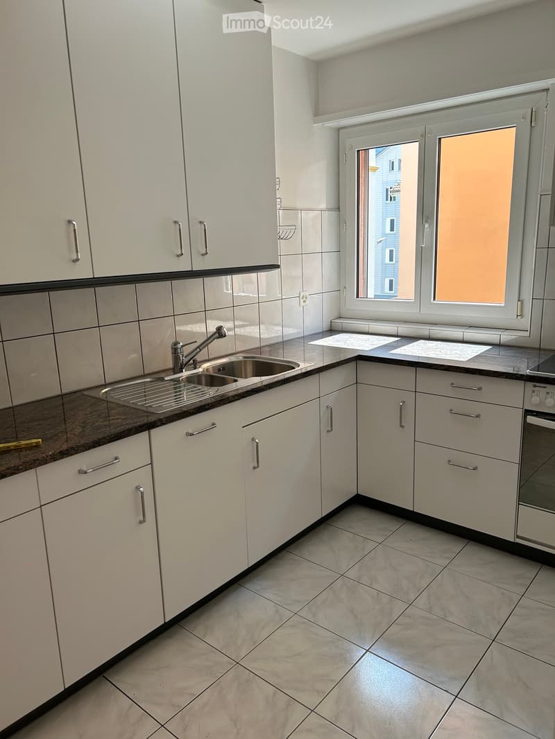 Modern kitchen with white cabinets, dark countertops, and a stainless steel sink. Features include a window with blinds and a tiled floor.
