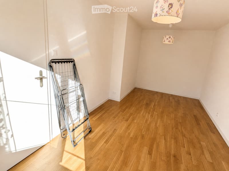 Empty room with wooden flooring, white walls, and a single door. A metal clothes drying rack is positioned near the door, and two hanging lamps are visible on the ceiling.